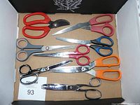 Photo showing eight assorted scissors with different handle colors and varying blade styles laid out on cardboard inside a box.