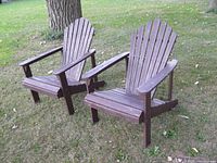 Two used pine Muskoka chairs placed on grass, showing front and left side views, detailing armrests and slatted design with some paint chipping.
