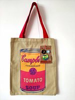 Full front view of beige canvas tote bag with red straps and Campbell's Tomato Soup can silkscreen print, displaying the attached kraft hangtag with three pins.
