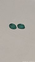 Two oval cut natural emeralds on a light background, showing their green color and polished surfaces.