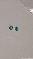 Pair of small oval cut emeralds placed on a white surface showing their size and color.