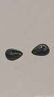 Close-up photo of the pair of pear-shaped blue-green sapphires on a plain background, showing color and shape.