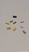 Ten small baguette cut sapphires of various colors scattered against a plain background.
