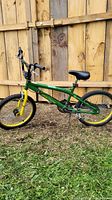 Side view of the 20-inch John Deere BMX style bicycle showing green and yellow frame, black seat, and tires on grass in front of a wooden fence.