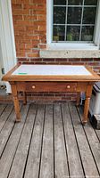 Front full view of the antique farmhouse table showing tile inlay top, drawer front, and wooden legs with casters.