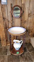 Full view of the antique wooden washstand featuring an oval mirror mounted between two vertical posts and a white ceramic basin seated on top. The washstand has four nicely turned twisted legs and a lower wooden platform holding the ceramic pitcher.