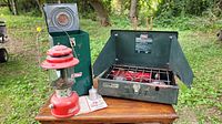Full view of Coleman lantern and stove on table outdoors with greenery in background.