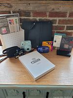 Lot contents on table showing cameras, gaming device, memory card, smart switch box, Adventure Challenge book, storage bin, and satellite receiver