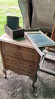 Small wooden storage chest with decorative front, three drawers, and damaged feet. Old metal washboard, rustic chalkboard and small wooden box placed on and near the chest.