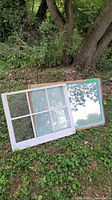 Antique 6-pane window with white distressed frame leaning against a large wooden framed mirror outdoors on grass surrounded by trees.