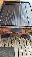 Small black metal patio bistro set with two chairs and one small table, showing entire set outdoors on a wooden deck with a brick wall and metal shutter background.