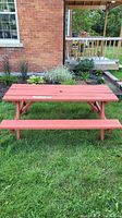Front view of freshly painted red wooden picnic bench with attached benches, positioned on grass by a brick wall and garden.