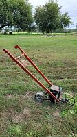 Full side view of the man powered garden planter showing the long orange wooden handles, metal seed hopper, wheel, and gearing mechanism on grassy field.