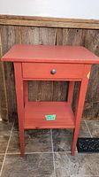 Front view of small red wood end table with one drawer and a lower shelf on tiled floor against wood paneling background.