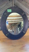Front view of the oval wall mirror showing the dark faux leather border with four brass belt buckles and the reflective mirror surface.