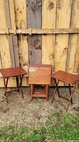 Seven collapsible wooden TV tray tables displayed outdoors against wooden fence, two tables set up with flat tops showing and five stacked folded.