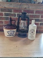 The full lot showing the Jack Daniel jug, Beacon Lantern, and Hudson's Bay whiskey bottle side by side on a wooden surface with brick wall background.