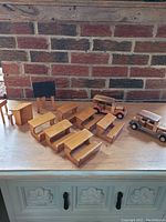 Entire set of vintage handmade wooden dollhouse school furniture including desks, chairs, chalkboard, and two handcrafted wooden cars on a surface against a brick wall.