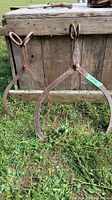 Two pairs of vintage metal ice tongs shown together on grass in front of a weathered wooden surface. The smaller pair is on the left, the larger on the right.