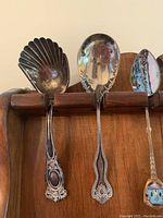 Close-up of three collector spoons on top row of wooden rack: one shell-shaped bowl spoon, one scalloped bowl spoon, one painted bowl spoon