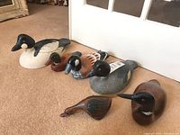Photo showing all six wooden waterfowl decoys arranged in a row on carpet floor, displaying variety in species and colors.