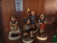 Five Hummel figurines displayed together on a wooden shelf, showing different children and animals.