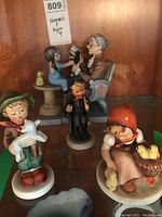 Four figurines including three Hummels and one unmarked depicting elderly woman reading to a girl