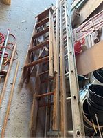 Photo showing two wooden ladders lying horizontally on a concrete floor with other stored items. The ladders show visible age and wear.