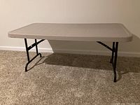 Front view of beige folding table set up, showing plastic top and black legs.