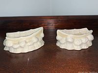 Two cream colored Belleek footed ashtrays shaped like small boats with floral detail along the sides and textured boat plank interior top rim. Displayed on dark wood surface.