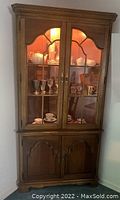 Full view of wooden corner cabinet with glass upper doors showing displayed tea sets and glassware, lower wooden doors closed.
