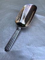 Silver plated ice scoop full view showing scoop and handle on gray textured fabric surface.