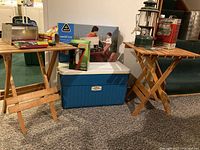 Wide view showing two wooden folding tables, large blue cooler, Coleman lantern on a table, and other camping items