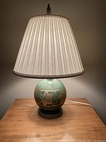 Photo showing the entire lamp including the base, lamp globe with green glaze and gold leaf floral design, pleated light beige fabric shade and tall finial at top.