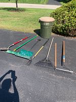 Full view of the long handled lawn and garden tools along with Rubbermaid Roughneck debris can outside on pavement.