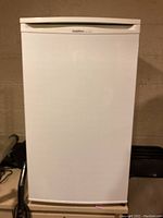 Front view of white mini refrigerator freezer closed, showing a plain white door and a handle integrated into the top edge of the door.