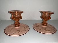 Pair of pink depression glass candlestick holders with etched leaf decoration on their bases, shown from a side angle.