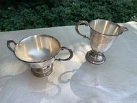 Two-piece silver set including a sugar bowl with two handles and a cream pitcher with one handle and spout, displayed on a white surface outside.