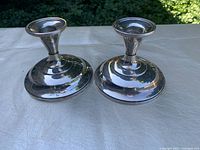 Pair of polished sterling silver weighted candlestick holders shown side by side on a white textured surface outdoors with natural light and greenery in background.