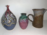 Photo showing all three pottery items: a uniquely shaped candle holder with flame cutouts in blue and red glaze, a small purple-green vase, and a speckled pitcher with handle.