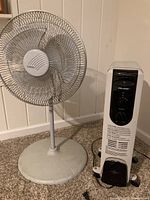 Pelonis space heater and Lasko oscillating floor fan positioned side by side against a wall on carpet.