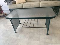 Full view of rectangular metal cocktail table with green frame, mesh side panels, textured glass top and metal rack underneath, photographed indoors on tiled floor with sofa in background.