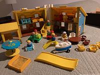 Overall view showing fold-out Fisher Price Family House open with interior graphics and multiple vintage Little People figures, playground accessories including blue umbrella table, yellow slide, sandbox, and several vehicles.