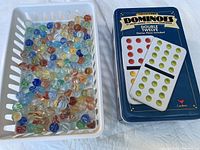 Top view of white basket filled with assorted glass marbles next to blue Cardinal dominoes tin box with double twelve set.