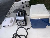 White electric egg poacher, black and silver Black & Decker toaster, and blue and white Igloo cooler on white folding table outdoors.
