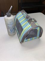 Pet carrier with blue, green, and yellow striped fabric, mesh side panel, and shoulder strap next to plastic water bottle with black spout.