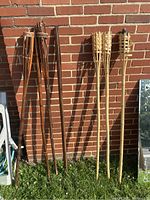 Eight wooden tiki torches leaning against a brick wall, six dark brown stained and two light natural wood torches with woven heads.