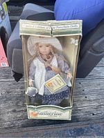 Front view of the vintage Katherine porcelain doll in original box, showing the doll with braided hair, white hat, vest, and floral skirt, along with the box labeling and some wear.