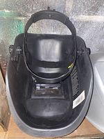Black welding helmet with adjustable headband and viewing window, shows used condition with some dirt and wear, photo taken to show condition and features.