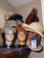 Photo showing three mannequin heads with painted facial details and various vintage ladies hats including a sequined turban, mustard yellow velvet hat, striped yellow and white hat, brown hat, and floral straw hat.